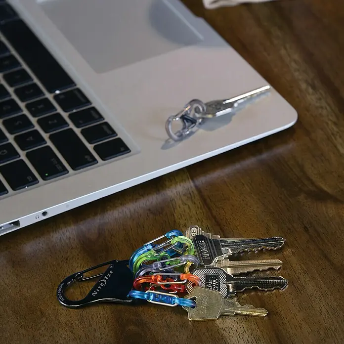 Nite Ize Mosquetón KeyRack - L x An: 12 x 2 mm, Acero inoxidable - Primer plano de un llavero Nite Ize con varias llaves en un escritorio junto a un portátil.