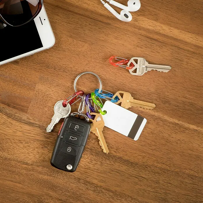 Nite Ize Karabinerhaken S-Biner KeyRing - L x B: 57 x 14 mm, Edelstahl - Schlüsselbund mit Autoschlüssel, Hausschlüsseln und Anhängern auf einem Holztisch, neben Handy und Kopfhörern.