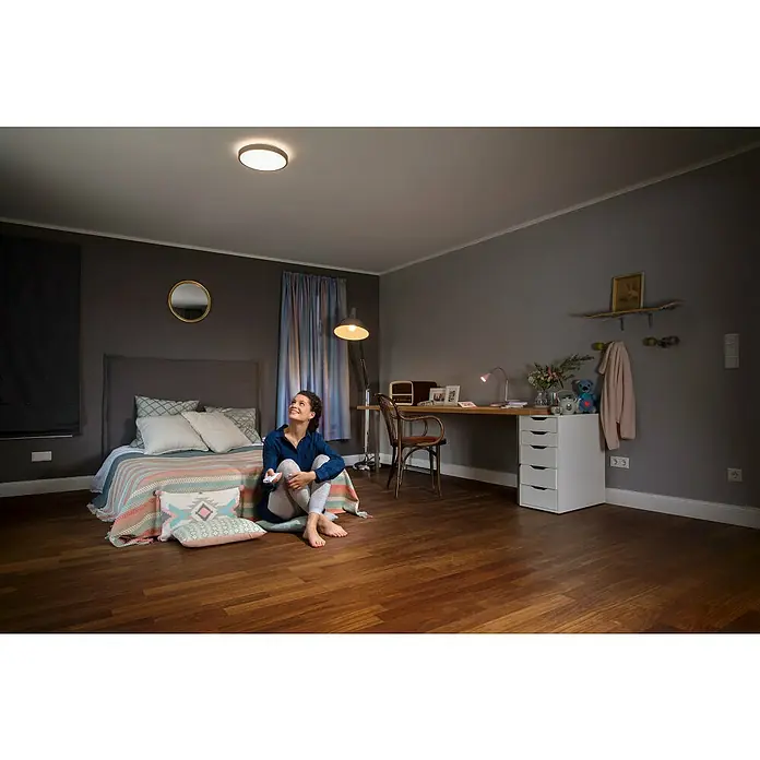 Ledvance LED-Deckenleuchte rund Silara - 21 W, Ø x H: 400 x 88 mm, Silber, Weiß, Neutralweiß - Eine Frau sitzt im Zimmer und blickt auf die Deckenleuchte, die ein warmes Licht ausstrahlt.
