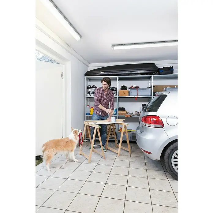 Ledvance LED-Feuchtraumlichtleiste Submarine - Länge: 60 cm, Lichtfarbe: Neutralweiß, 12 W, IP65 - Ein Mann streicht Holz auf einer Werkbank in einer Garage, während ein Hund mit einer Leine daneben steht.