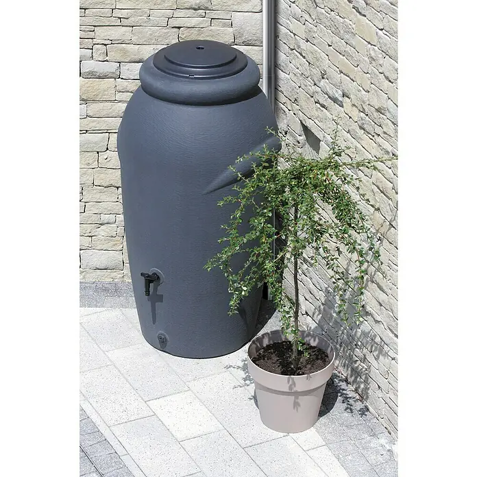 Regenspeicher Aquacan Baby - 210 l, Grau - Graue Regentonne aus Kunststoff mit Hahn und Deckel, neben einer Pflanze auf einer gepflasterten Fläche an einer Steinmauer.