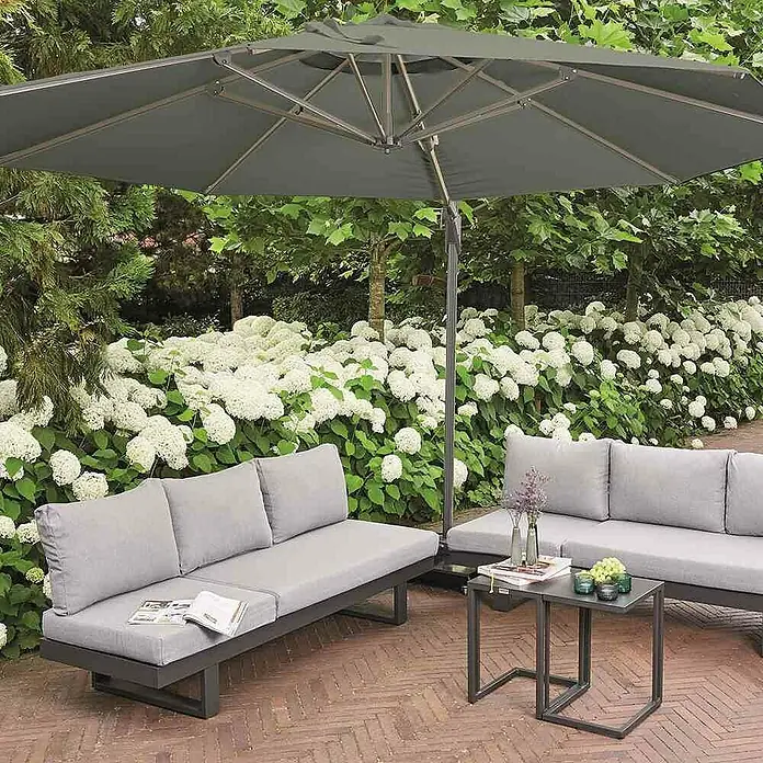 Siena Garden Ampelschirm Sunset N+ - Olive, Bespannungsmaß (L x B): 350 x 350 cm - Eine Gartensitzgruppe mit grauen Polstern und einem Sonnenschirm, umgeben von üppigen Hortensienbüschen für entspannte Stunden im Freien.