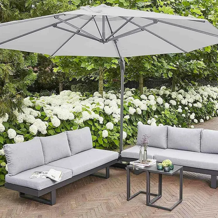 Siena Garden Ampelschirm Sunset N+ - Stone, Bespannungsmaß (L x B): 350 x 350 cm - Eine stilvolle graue Terrassenlounge mit Sonnenschirm und Couchtisch, umgeben von üppiger grüner Vegetation und weißen Hortensien.