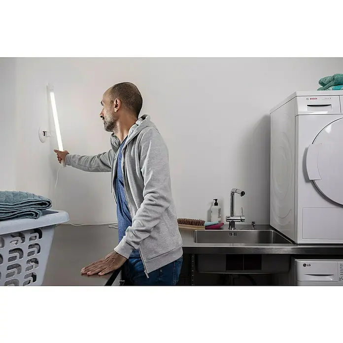 Osram LED-Unterbauleuchte TubeKIT - 19 W, Länge: 1.200 mm, Neutralweiß - Ein Mann schaltet in einem Waschraum eine schmale LED-Wandleuchte ein, neben Waschmaschine, Spüle und einem Wäschekorb mit Textilien.