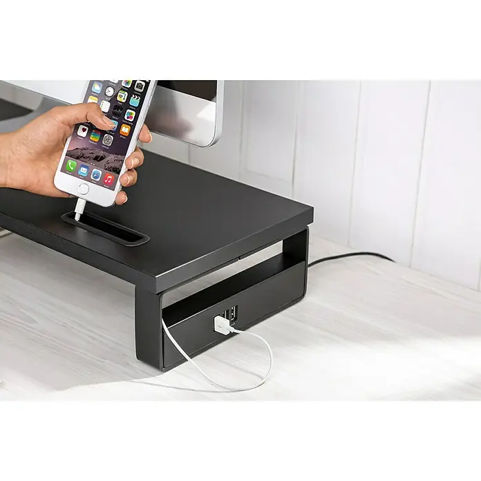 Voltomat Monitor-Erhöhung - Schwarz - Nahaufnahme eines schwarzen Monitorständers mit USB-Anschlüssen, an dem ein Smartphone angeschlossen ist, auf einem Schreibtisch.