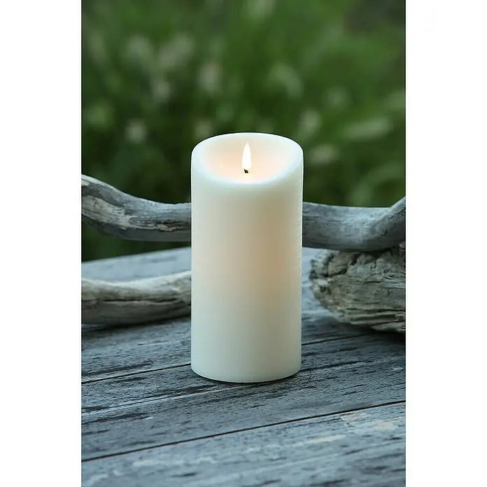 Vela LED para exteriores Elephant - Ø x Al: 10 x 20 cm, Crema, Cera natural - Primer plano de una vela de pilar color crema con luz cálida, colocada sobre una mesa de madera rústica contra un fondo verde borroso, ideal para el ambiente y la decoración.