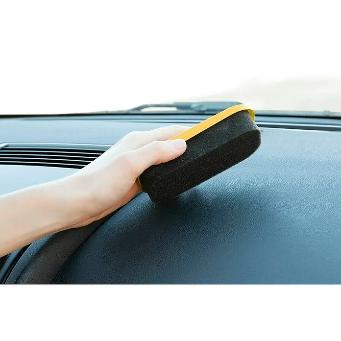 Esponja limpiadora para salpicaderos - Vainilla - La imagen muestra una mano usando una esponja amarilla y negra para limpiar el tablero de un coche, destacando el producto de limpieza.