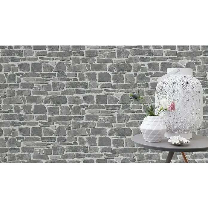 Rasch Papiertapete - Grau, Steinoptik, 10,05 x 0,53 m - Das Bild zeigt ein weißes Deko-Objekt und eine Vase mit Blumen auf einem grauen Tisch vor einer grauen Steinwand.