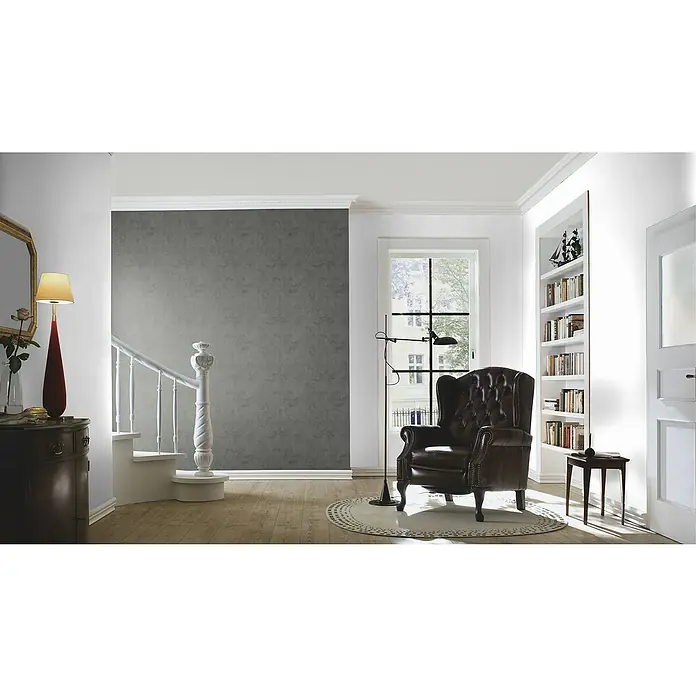 Rasch Tapetenwechsel Vliestapete - Dunkelgrau, Betonoptik, 10,05 x 0,53 m - Gemütliches Interieur mit einem braunen Ledersessel, einer Stehlampe und einem Bücherregal in einem hellen Raum mit Holzboden.