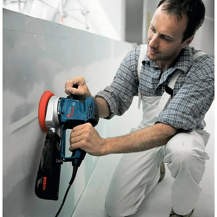 Bosch Professional Elektro-Exzenterschleifer GEX 34-150 - 340 W, Durchmesser Schleifteller: 150 mm, Leerlaufdrehzahl: 4.500 U/min - 12.000 U/min - Ein Handwerker benutzt einen Bosch Wandschleifer zum Glätten einer Wand. Der Schleifer ist blau und hat einen roten Schleifkopf.