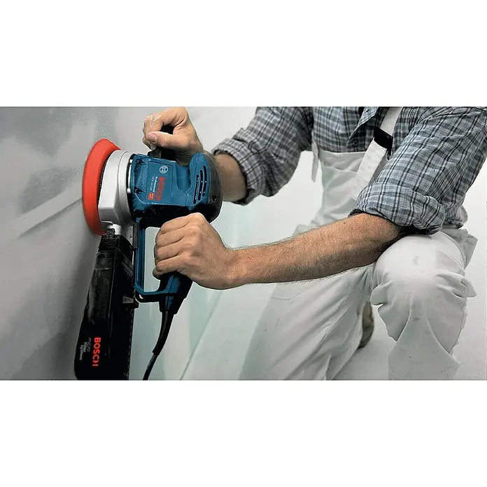 Bosch Professional Elektro-Exzenterschleifer GEX 34-150 - 340 W, Durchmesser Schleifteller: 150 mm, Leerlaufdrehzahl: 4.500 U/min - 12.000 U/min - Ein Handwerker glättet eine Wand mit einem blauen Bosch Wandschleifer mit Staubabsaugung für ein sauberes Arbeitsumfeld. Präzise und effiziente Oberflächenbearbeitung.
