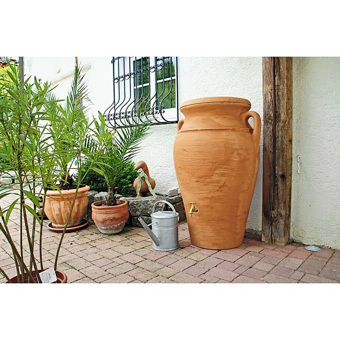 Arves Regenspeicher Amphore Helena - 300 l, Terra/Antik - Ein großer Terrakotta-Wasserspeicher mit einem Messinghahn steht auf einer gepflasterten Terrasse, neben Pflanzen und einer Gießkanne.
