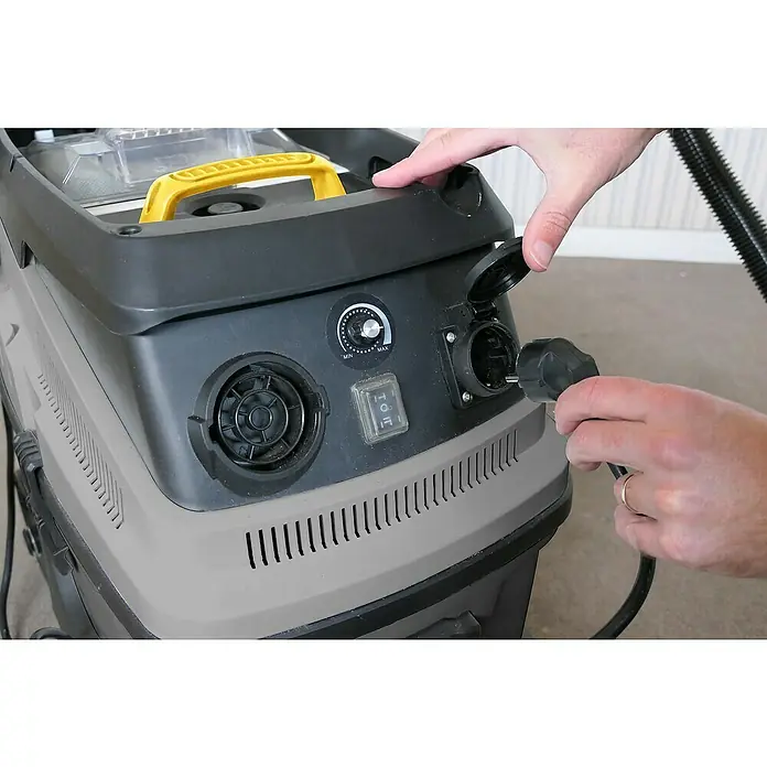 Fartools Aspirador en seco y húmedo eléctrico MF35 - 1.600 W, Volumen de recipiente: 35 l, Con bolsa - Un hombre conecta un cable de alimentación negro a una aspiradora gris y negra con asa amarilla.