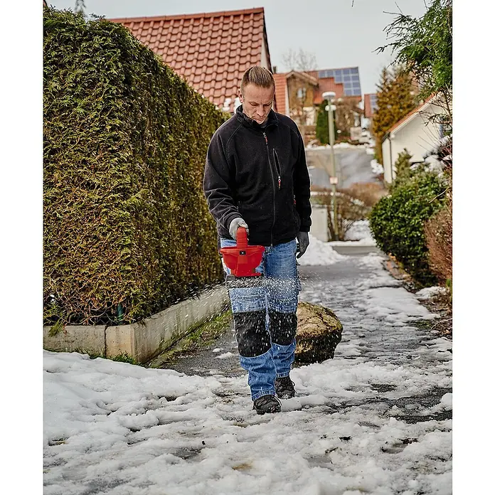Einhell Power X-Change 18V Handstreuer GE-US 18 Li-Solo - Streubreite: 5 m, Fassungsvermögen: 3,3 l - Mann streut Salz mit einem roten Salzstreuer auf einen vereisten Gehweg, um die Sicherheit zu erhöhen. Effektive Eisbekämpfung mit Streusalz.