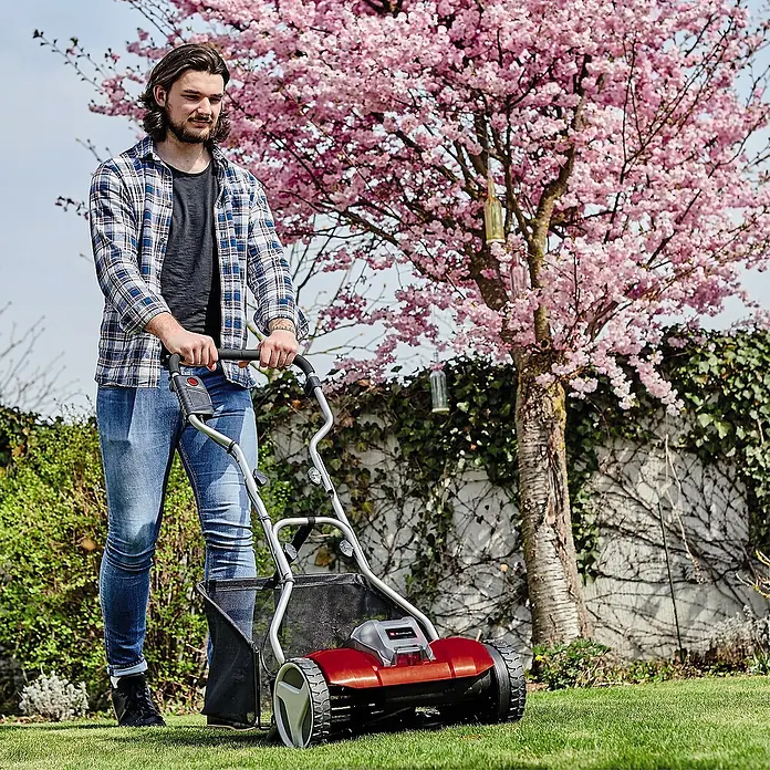 Einhell Power X-Change 18V Akku-Spindelmäher GE-HM 18/38 Li-Solo - 18 V, Ohne Akku, 4 Ah, Schnittbreite: 38 cm - Ein Mann benutzt einen Einhell Rasenlüfter im Garten, umgeben von blühenden Bäumen und Grün.