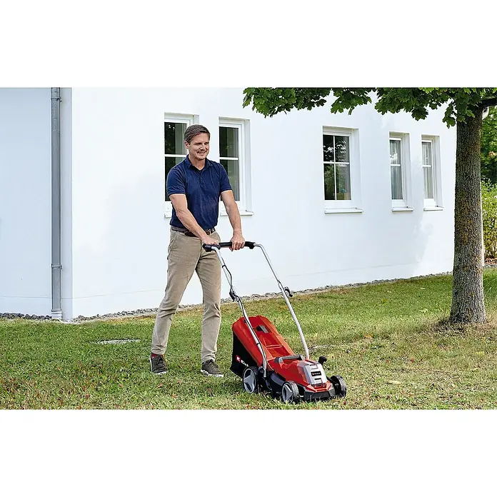 Einhell PXC 18V Cortacésped de batería GE-CM 18/33 Li - 18 V, 1 batería, 4 Ah, Ancho de corte: 33 cm - Un hombre está cortando el césped con una cortadora de césped Einhell roja frente a una casa blanca.