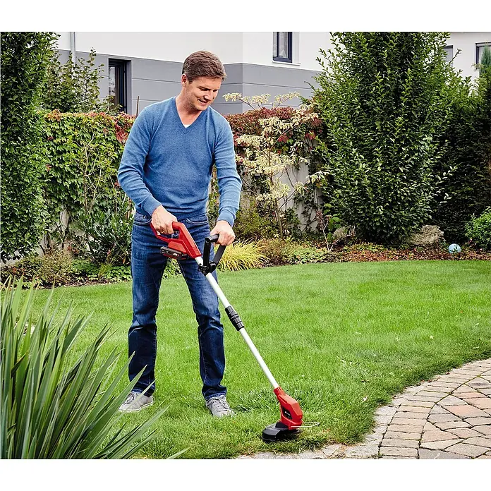 Einhell Power X-Change 18V Akku-Rasentrimmer - 18 V, 2 Ah, Li-Ionen, 1 Akku, Schnittbreite Messer: 24 cm - Ein Mann benutzt einen Rasentrimmer, um das Gras zu schneiden, das an einem gepflasterten Weg in seinem Garten wächst.