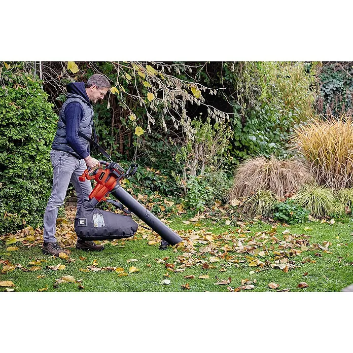 Einhell PXC 18V Soplador/Aspirador de batería 36/230 LI E-SOLO - 18 V, Sin batería, Potencia de soplado: 225 m³/h, Velocidad de soplado: 230 km/h - Un hombre usa un soplador de hojas Einhell rojo con una bolsa de recogida negra para limpiar hojas en un césped verde.