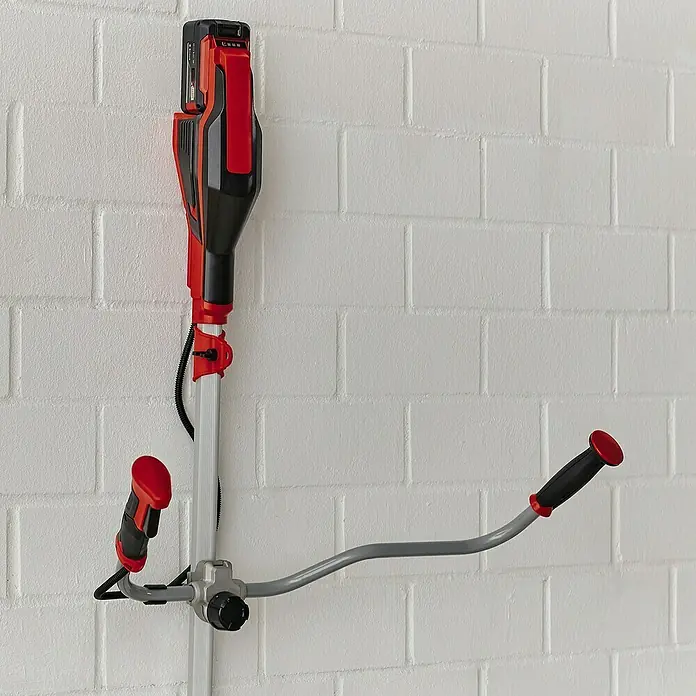 Einhell PXC 18V Desbrozadora de batería Agillo - 18 V, Iones de litio, Sin batería - La imagen muestra un cortacésped inalámbrico rojo y negro montado en una pared de ladrillo blanco, mostrando su diseño de almacenamiento compacto.