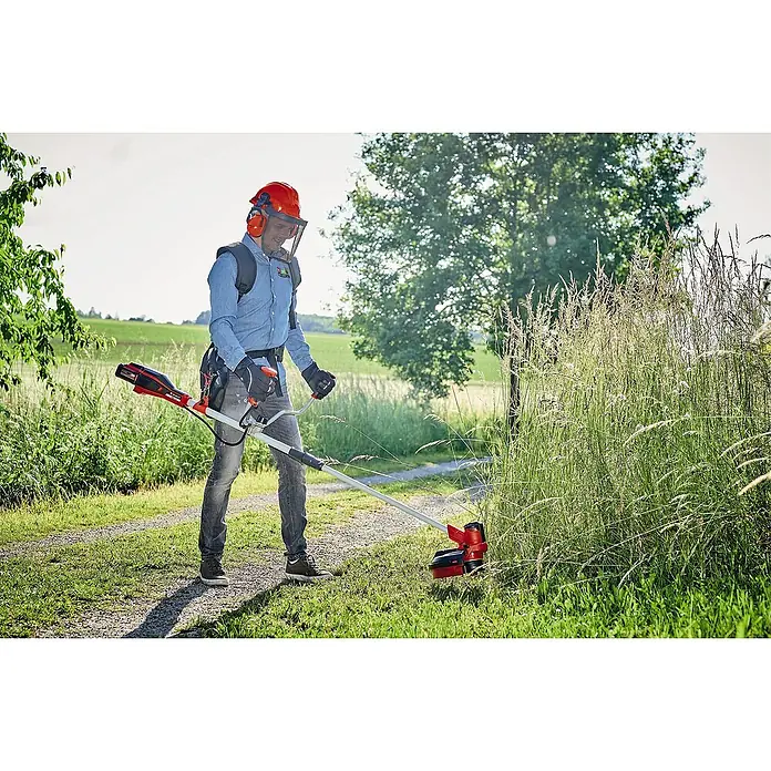 Einhell PXC 18V Desbrozadora de batería Agillo - 18 V, Iones de litio, Sin batería - Un hombre está cortando el césped con una desbrozadora. La desbrozadora es roja y negra, y está siendo utilizada por una persona con equipo de seguridad.