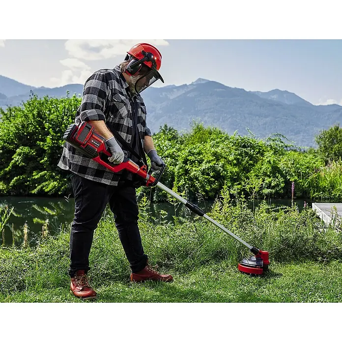 Einhell PXC 18V Multiherramienta de jardín 36VGE-LM 36/4in1 + Batería y cargador PXC-Starter kit - 36 V, Iones de litio, 4 Ah, 2 baterías - Un hombre trabajando en el jardín con un cortacésped inalámbrico Einhell rojo, con montañas al fondo.