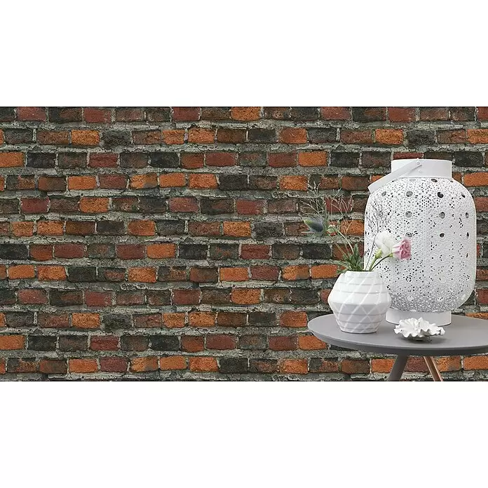 Rasch Factory 4 Vliestapete Steinmauer - Rotbraun, Steinoptik, 10,05 x 0,53 m - Weiße Dekoobjekte: eine verzierte Laterne, eine geometrische Vase mit Blumen und eine Muschel auf einem grauen Beistelltisch.
