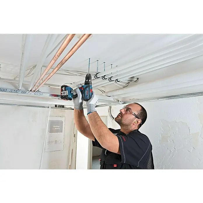 Bosch Professional AMPShare 18V Akku-Schlagbohrschrauber GSB 18V-28 - 18 V, 2 Akkus, 2 Ah - 4 Ah, 63 Nm, Inkl. Zubehörkoffer - Der Handwerker befestigt mit einer blauen Bosch Professional Akku-Bohrmaschine Rohre an einer Decke. Schutzbrille und Handschuhe inklusive.