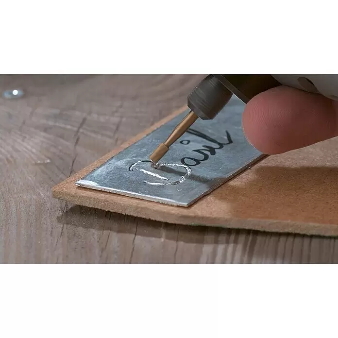 Dremel Fräsmesser Max 194DM - 2 Stk. - Eine Hand prägt mit einem bronzefarbenen Werkzeug silberne Metallbuchstaben "Basil" auf ein dunkelgraues Metallschild.