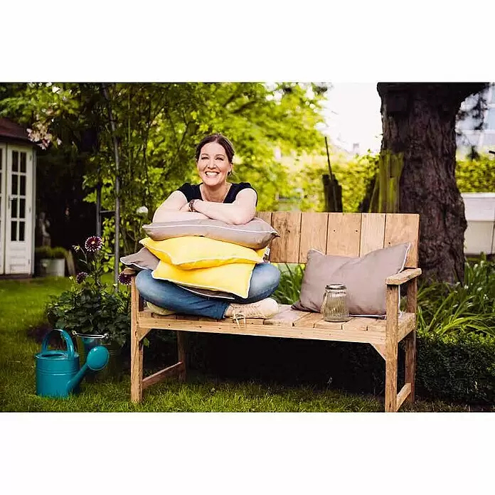 Siena Garden Gartenbank Loreo - B x T x H: 124 x 57 x 90 cm, Teak, Natur - Eine Frau sitzt lächelnd auf einer Holzgartenbank mit grauen und gelben Kissen.