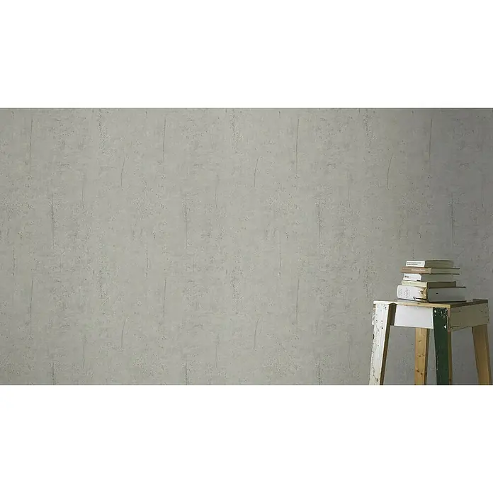 Rasch Vliestapete Beton - Hellgrau, Betonoptik, 10,05 x 0,53 m - Ein Stapel Bücher liegt auf einem kleinen, weiß-grünen Hocker vor einer grauen Wand, ideal für eine ruhige Leseecke.