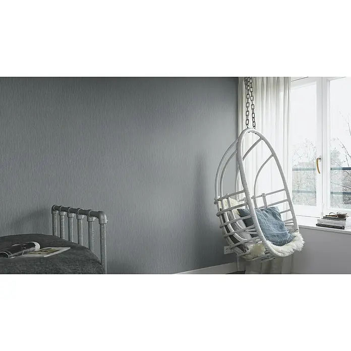 Rasch Berlin Vliestapete - Dunkelgrau, Uni, 10,05 x 0,53 m - Hängesessel mit Kissen und Decke neben einem Fenster mit Blick auf eine verschneite Landschaft. Der Hängesessel ist aus weißem Rattan gefertigt.