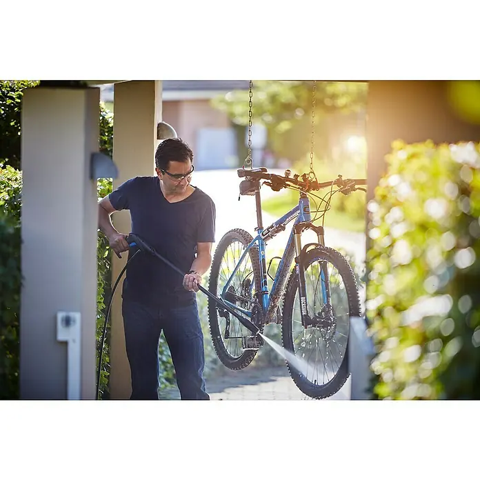 Nilfisk Hochdruckreiniger C 135.7-6 Garden X-tra - 1.500 W, Max. Druck: 135 bar, Fördermenge: 460 l/h - Ein Mann reinigt ein blaues Mountainbike mit einem Hochdruckreiniger, das an Ketten hängt.