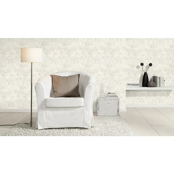 Rasch Vliestapete Beton - Altweiß, Betonoptik, 10,05 x 0,53 m - Das Bild zeigt ein stilvolles Wohnzimmer mit einem weißen Sessel, einer Stehlampe und einem Regal mit Dekorationselementen. Die Tapete hat eine dezente Struktur.