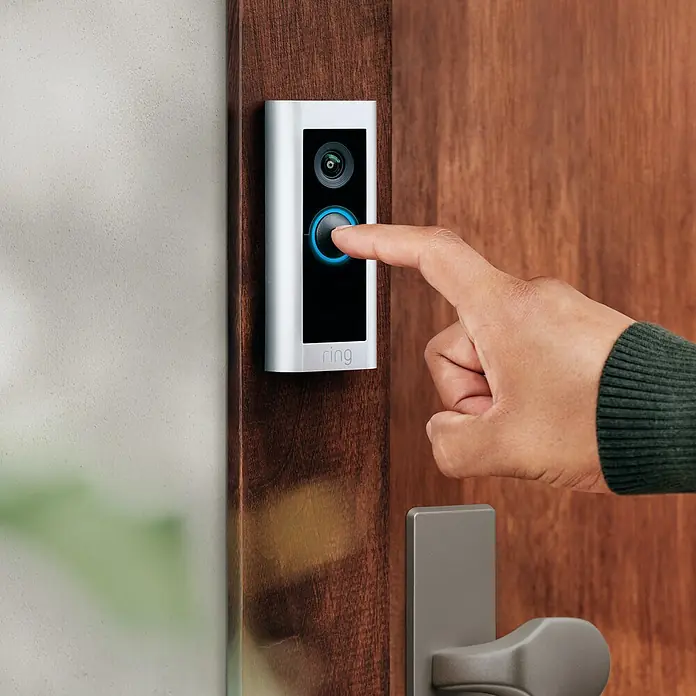 Ring Türklingel mit Kamera Video Doorbell Pro 2 Plug-in - Netzanschluss, Nickel matt, 1536p HD, 2,2 x 4,9 x 11,4 cm, Smarte Steuerung: Ring App - Finger drückt auf die Klingeltaste eines Ring-Video-Türsprechers, montiert an einer Holztür.