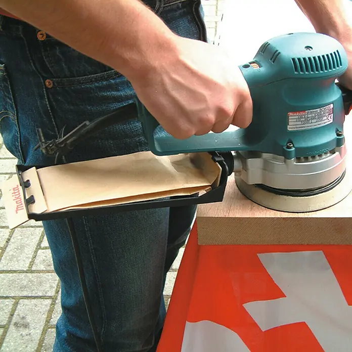 Makita Elektro-Exzenterschleifer BO6030JX - 310 W, Durchmesser Schleifteller: 150 mm - Nahaufnahme eines Makita Exzenterschleifers, der von einer Person gehalten wird, mit Blick auf den Staubbeutel und die Bedienelemente.