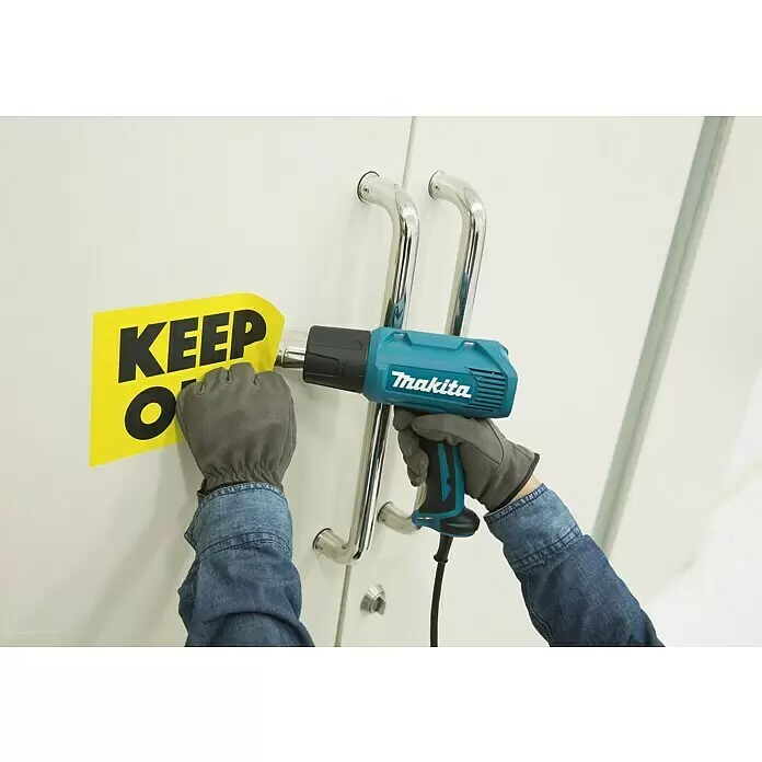Makita Heißluftgebläse HG5030K - 1.600 W, Temperaturbereich: 350 °C - 500 °C - Eine Person entfernt mit einem türkis-schwarzen Makita Heißluftgebläse einen gelben 'KEEP OUT'-Aufkleber von einer weißen Tür.