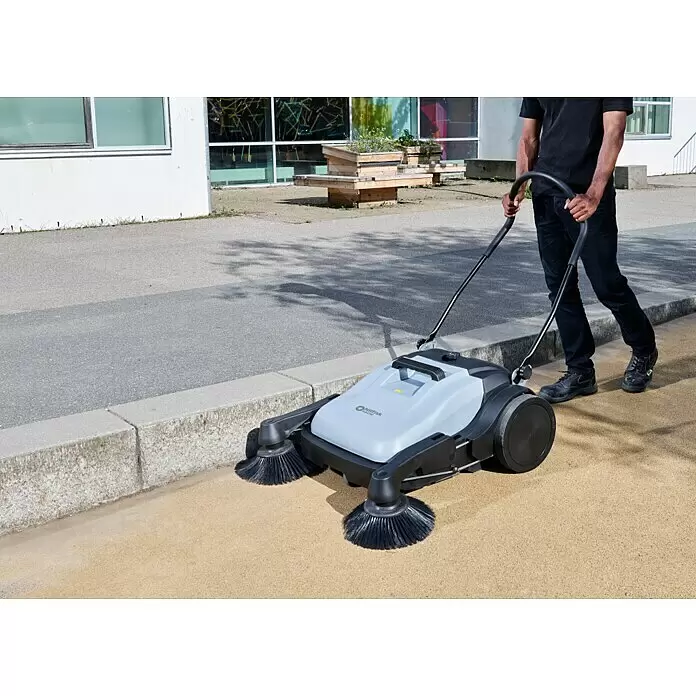 Nilfisk Handkehrmaschine SW 250 - Flächenleistung: 1.400 m²/h - 1.840 m²/h, Behältervolumen: 38 l - Mann schiebt eine graue und schwarze Kehrmaschine mit zwei Frontbürsten auf einem Außenweg.
