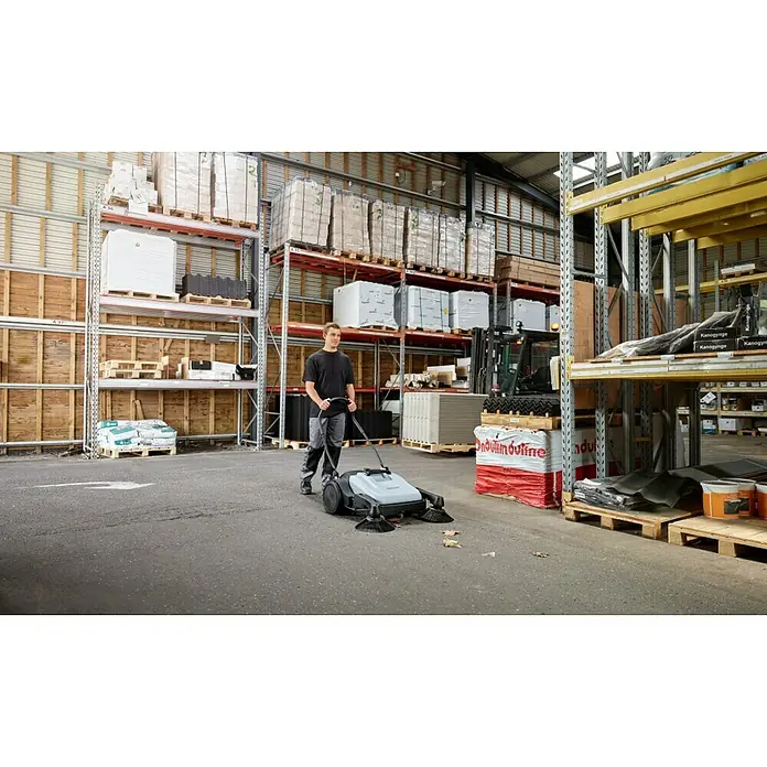 Nilfisk Handkehrmaschine SW 250 - Flächenleistung: 1.400 m²/h - 1.840 m²/h, Behältervolumen: 38 l - Ein Mann bedient eine Kehrmaschine in einem Lagerhaus. Die Maschine reinigt effizient den Boden von Schmutz und Ablagerungen.