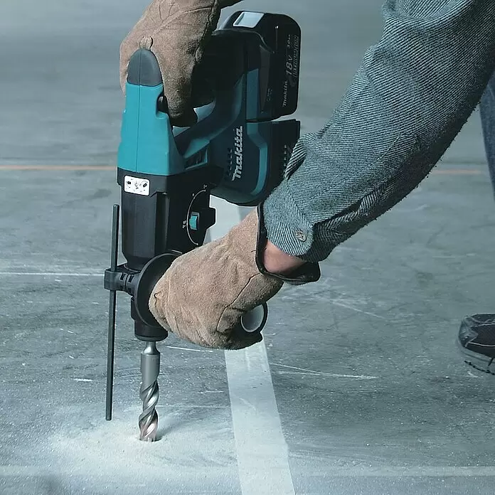 Makita LXT 18V Martillo combinado de batería DHR243RTJ - 18 V, 2 baterías, 5 Ah, 2 J, Con extracción de polvo - Persona usando un taladro percutor a batería Makita turquesa y negro con una broca grande para perforar un suelo de hormigón, creando polvo.