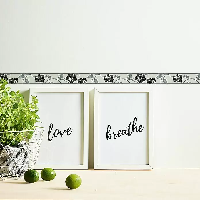 A.S. Création Only Borders 11 Tapetenborte - Kunststoff, Schwarz/Weiß - Dekorative Wandbordüre mit wiederholtem grauem Blumenmuster auf hellgrauem Hintergrund, angebracht an einer Wand über gerahmten Bildern und einer Pflanze.