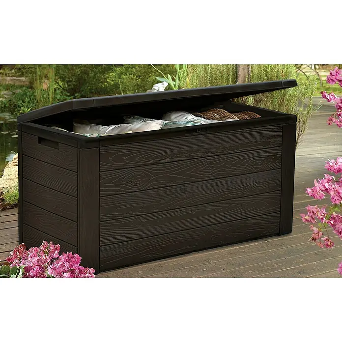 Jelenia Plast Arcón para cojines de exterior Woody - 119 x 45 x 57 cm, Plástico, Antracita - Una caja de almacenamiento de plástico marrón con aspecto de madera y tapa ligeramente abierta en un jardín.