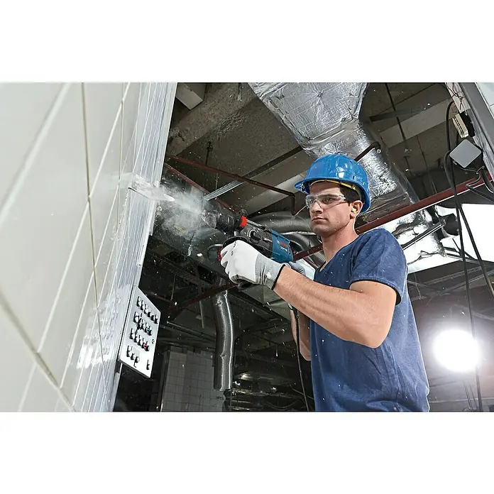Bosch Professional Kombihammer GBH 2-28 F - 880 W, 3,2 J, SDS-Plus-Aufnahme, Ohne Bohrer & Meißel - Ein Mann bohrt mit einer Bosch-Bohrmaschine ein Loch in eine geflieste Wand, wobei Staub aufgewirbelt wird. Er trägt Schutzkleidung.