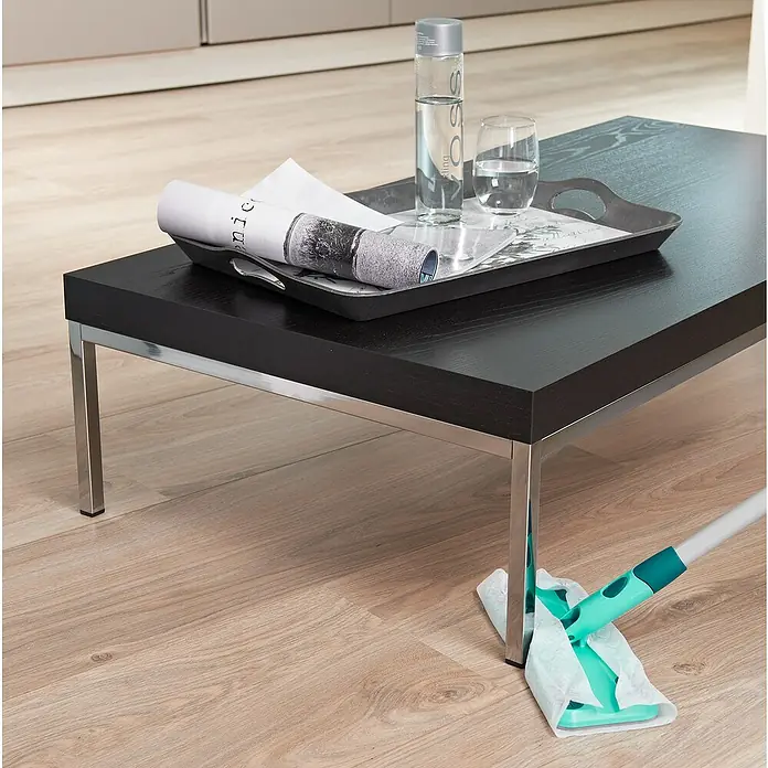 Leifheit Set de mopa atrapapolvos Clean & Away - Ancho: 22 cm - Imagen que muestra una mesa de centro negra con revistas, agua y una toallita de limpieza apoyada en una pata. El enfoque está en el proceso de limpieza.
