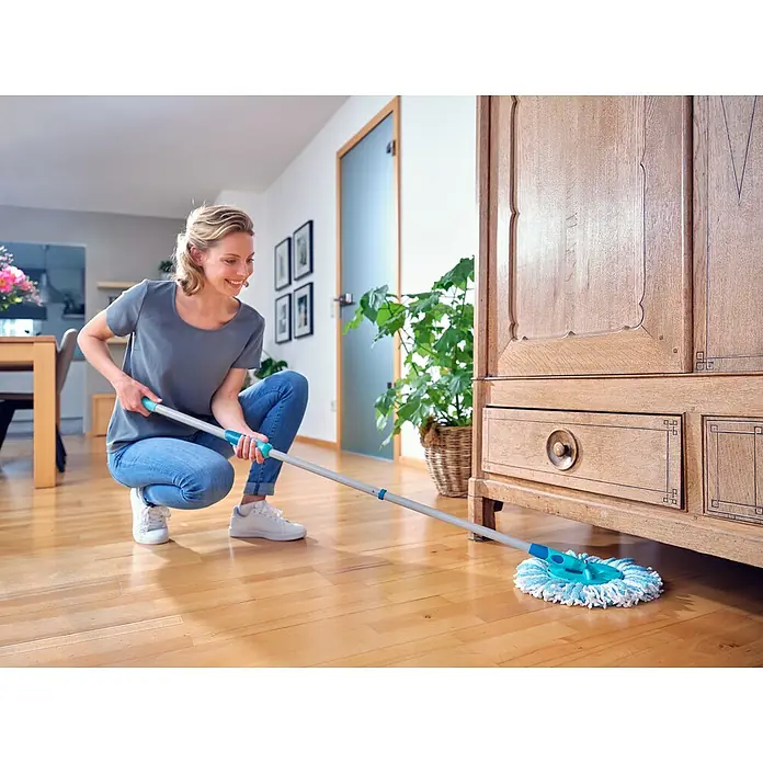 Leifheit Wischmop-Set CLEAN TWIST Disc Mop Ergo - 3 -tlg., 6 l, Grünblau - Frau wischt den Holzboden mit einem blauen Wischmopp mit Teleskopstiel unter einem Schrank. Der Boden wird sauber und glänzend.