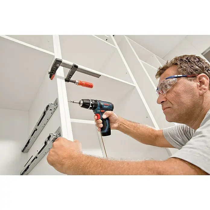 Bosch Professional Akku-Schlagbohrschrauber GSB 12V-15 - 12 V, 2 Akkus, 2 Ah - 4 Ah, 30 Nm, 2 Akkus + Ladegerät + 25-tlg. Bit-Set/14-tlg. Bohrer-Set + Tasche - Ein Mann befestigt Schubladenführungen mit einem Bosch Akku-Bohrschrauber an einem weißen Schrank. Sicherheitsbrille schützt seine Augen.