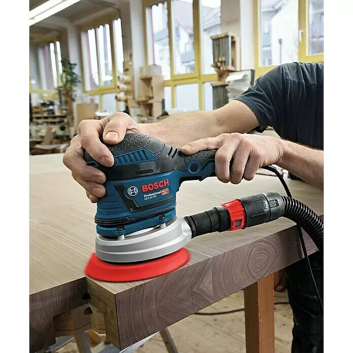 Bosch Professional Elektro-Exzenterschleifer GEX 40-150 - 400 W, Durchmesser Schleifteller: 150 mm - Eine Person verwendet einen blau-schwarzen Bosch Exzenterschleifer mit rotem Schleifteller zum Glätten einer Holzoberfläche. Ein Absaugschlauch ist angeschlossen.