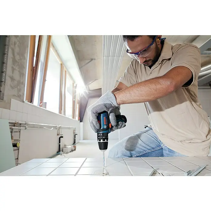 Bosch Professional Akku-Schlagbohrschrauber GSB 12V-15 - 12 V, 2 Akkus, 2 Ah - 4 Ah, 30 Nm, 2 Akkus + Ladegerät + 25-tlg. Bit-Set/14-tlg. Bohrer-Set + Tasche - Ein Handwerker bohrt mit einer blauen Bosch Akku-Bohrmaschine ein Loch in weiße Fliesen. Er trägt dabei Schutzbrille und Arbeitshandschuhe.