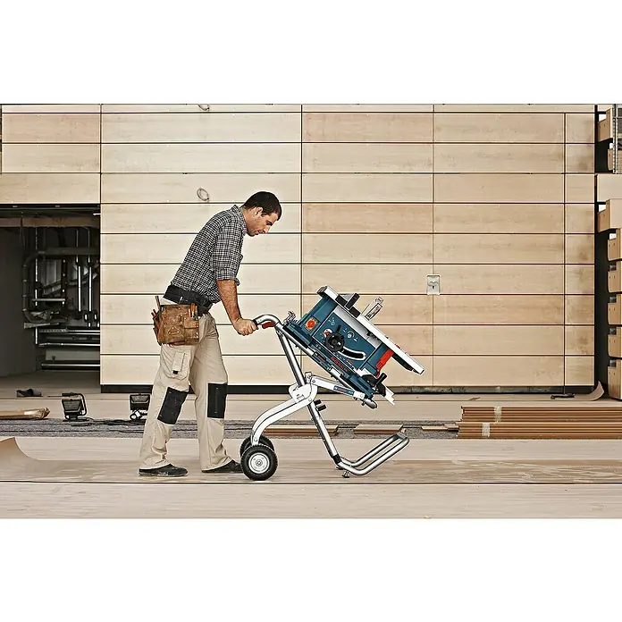 Bosch Professional Werkbank GTA 60 W - Arbeitshöhe: 622 mm, Belastbarkeit: 180 kg - Ein Bauarbeiter transportiert eine Bosch Tischkreissäge auf einem Rollwagen durch eine Baustelle. Das Gerät ist blau und weiß.