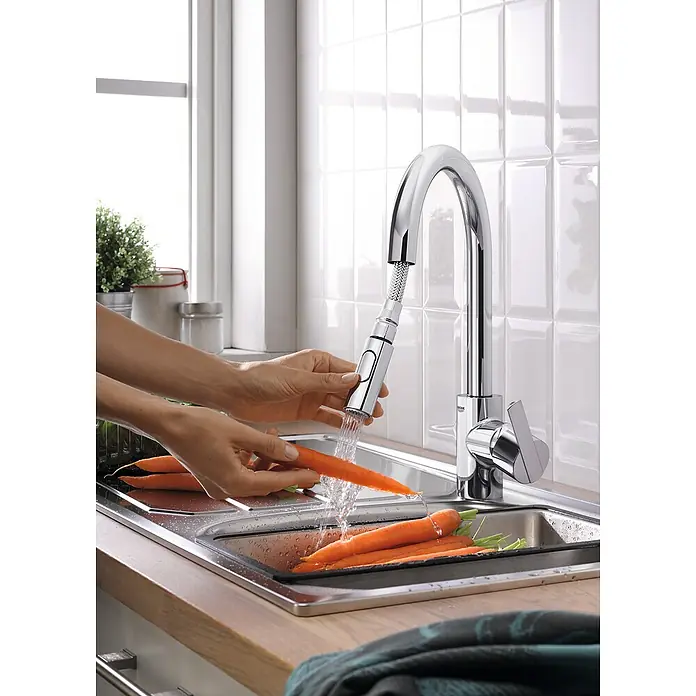 Grohe QuickFix Feel Grifo de cocina - Profundidad del grifo: 223 mm, Cromo, Brillante - Un brillante grifo de cocina Grohe con rociador extraíble lavando zanahorias, rodeado de azulejos y una ventana, mostrando un diseño moderno.