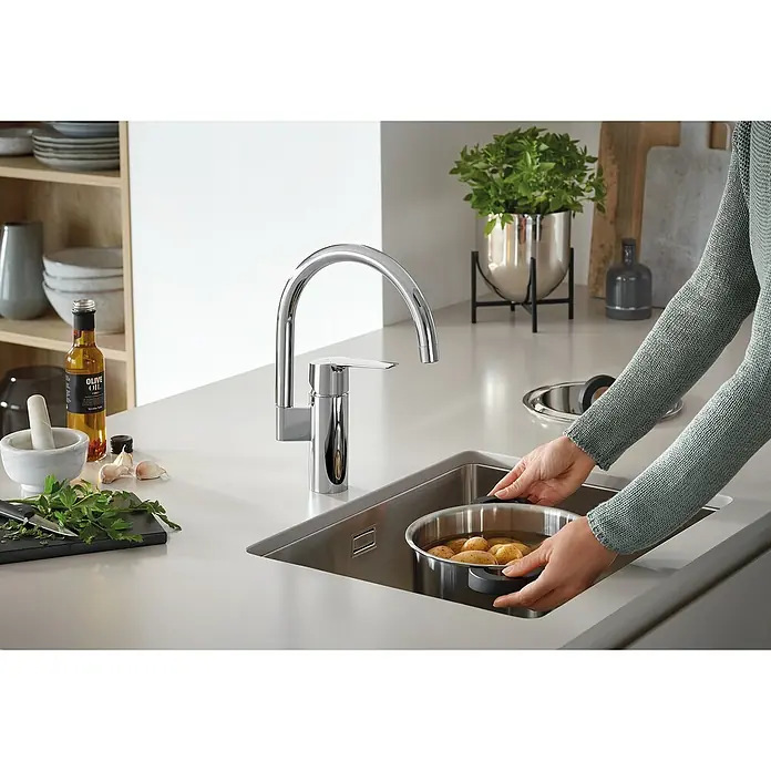 Grohe QuickFix Grifo de cocina Start 2021 - Profundidad del grifo: 183 mm, Cromo, Brillante - Un moderno grifo de cocina cromado es el centro de atención mientras una persona coloca una olla de papas en el fregadero. Diseño elegante y funcionalidad.